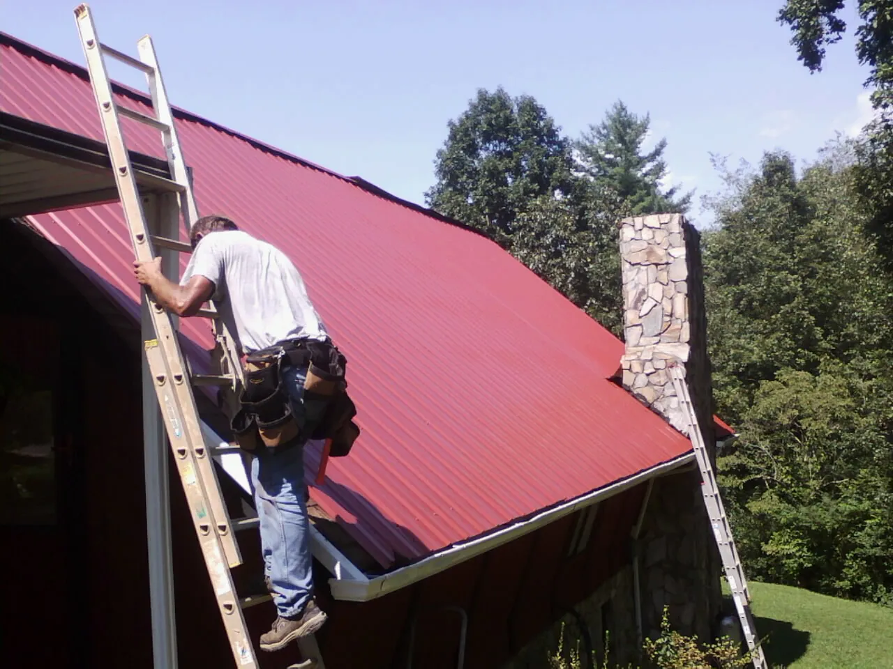 Roofer on a ladder beside a red metal roof and stone chimney — Soffit & Fascia Repair by Roofing Brothers in Salinas
